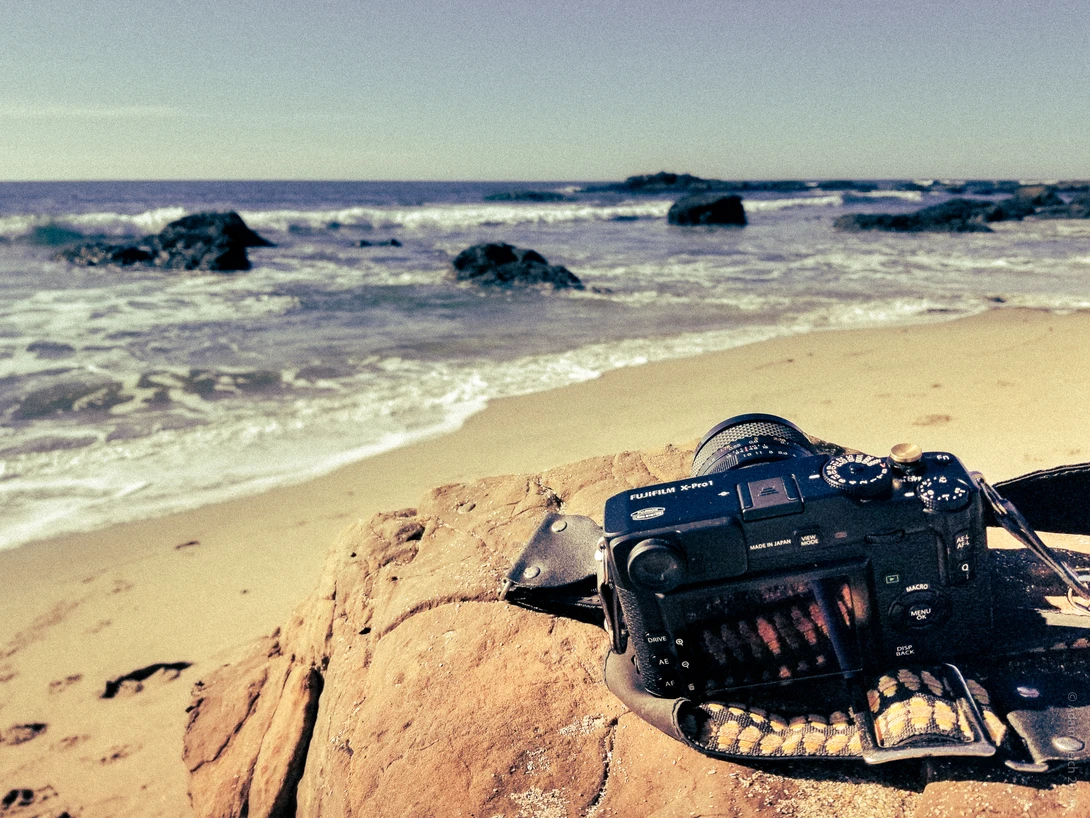 DSLR camera on sandy beach with ocean waves and rocks in background.
