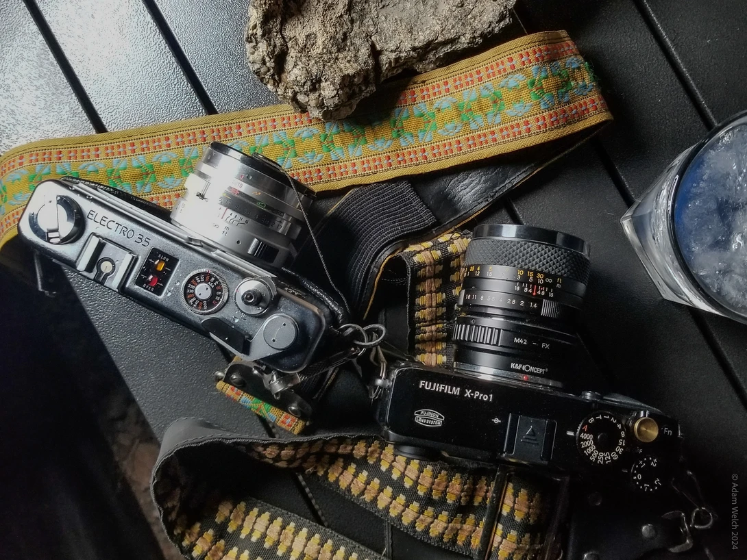 Vintage film cameras, lenses, and film rolls arranged on dark surface with decorative fabric trim.