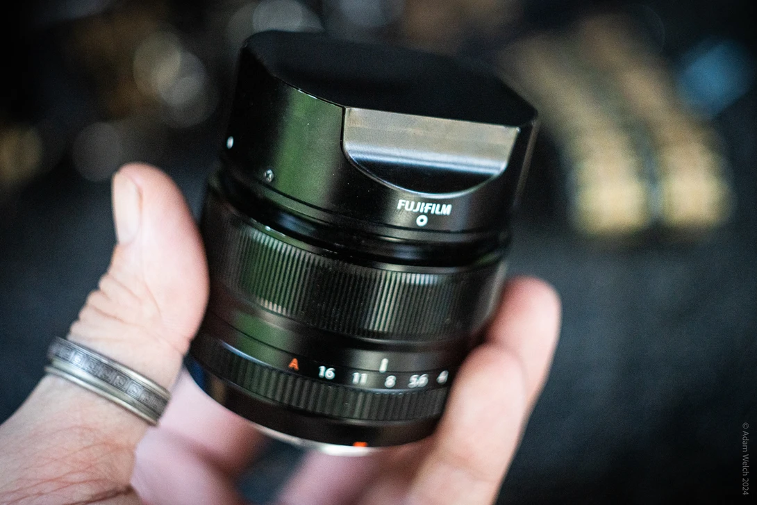 Hand holding a Fujinon prime lens with focus ring and aperture markings visible against blurred background.
