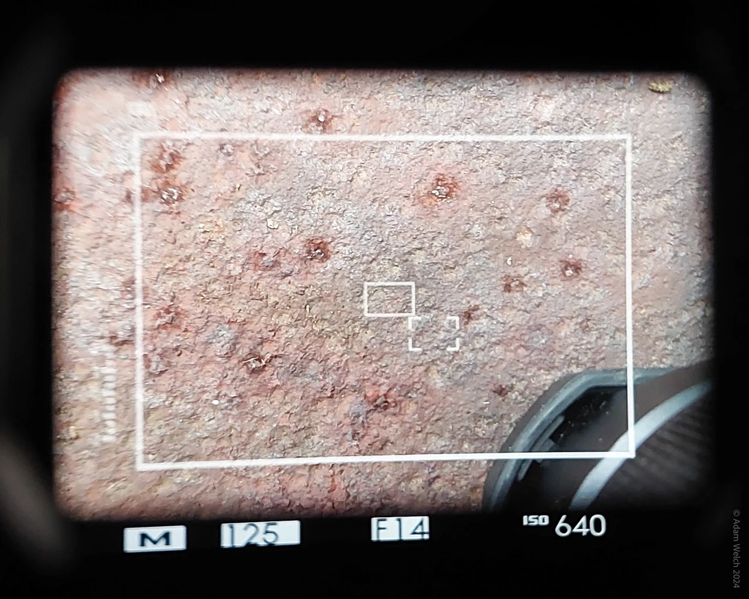 Through-the-lens view of desert ground with exposure metering grid overlay and camera settings display.
