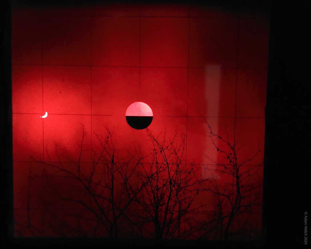 Red sun with dark horizontal band bisecting it, silhouetted bare branches in foreground against crimson sky.
