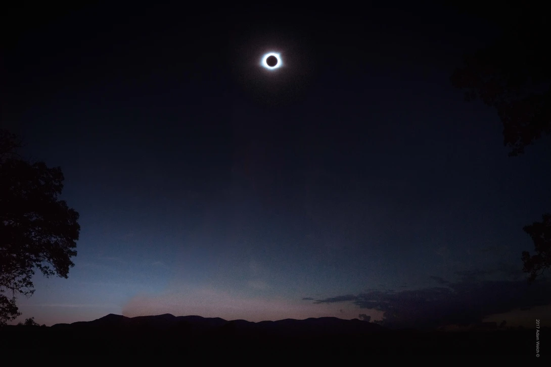 Total solar eclipse with corona visible against twilight sky above silhouetted landscape.