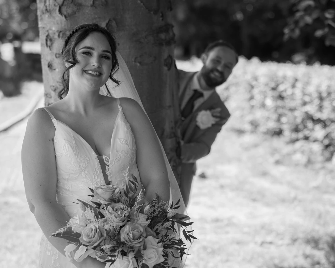Bride in white dress smiling at camera while groom peeks from behind tree in background.