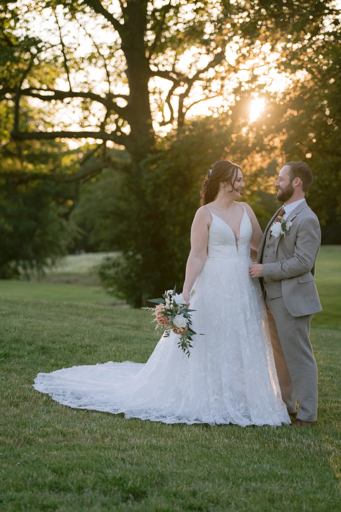 Bride and groom standing together on grass during golden hour, bride holding bouquet with long train visible.