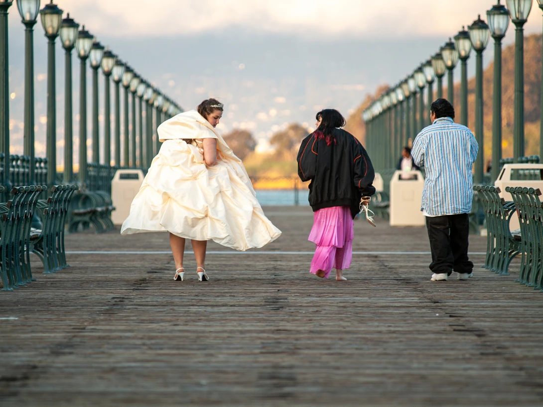 Bride in white dress walking with two people on a tree-lined pedestrian bridge at golden hour.