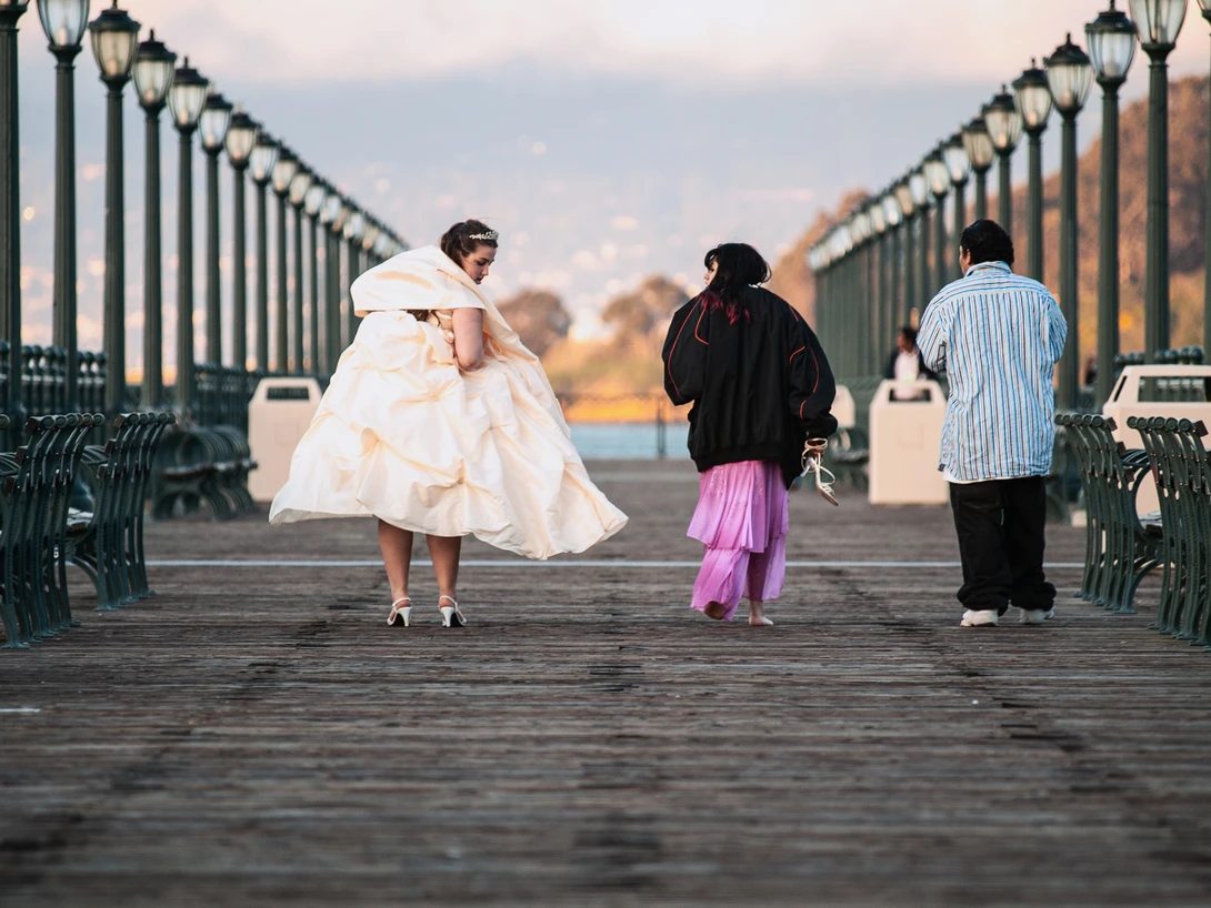 Three people walking on a wooden pier lined with lamp posts, shot from behind during golden hour.