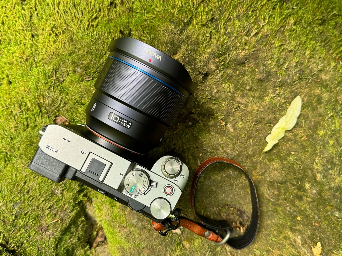 Mirrorless camera with attached lens on moss-covered ground next to a wrist strap.