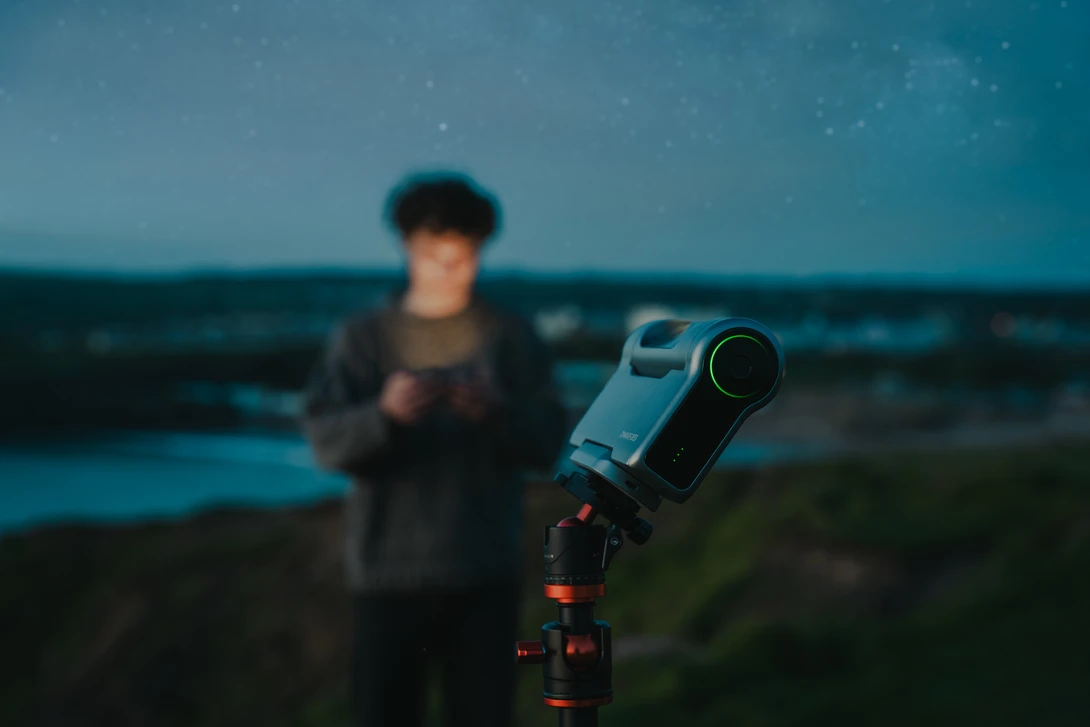 Telescope mounted on tripod pointed at twilight sky with blurred figure and landscape in background.