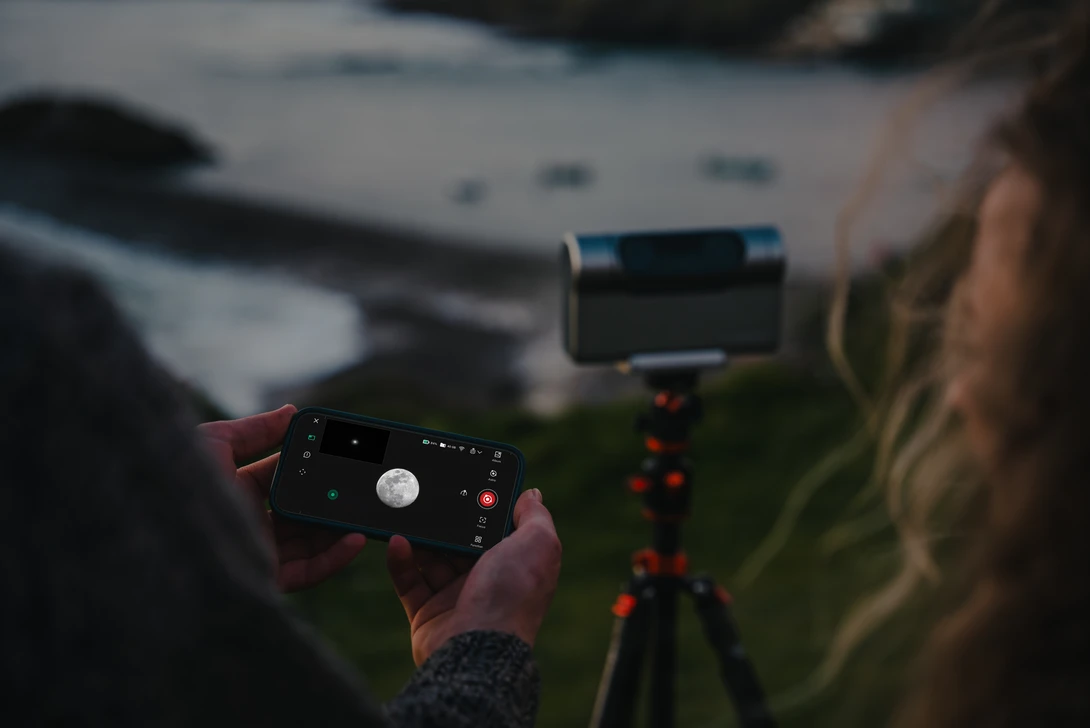 Photographer holding a smartphone on a tripod while operating a camera remote control outdoors.