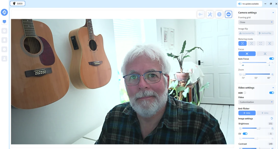 Man with white beard and glasses seated in front of two acoustic guitars hanging on a white wall.
