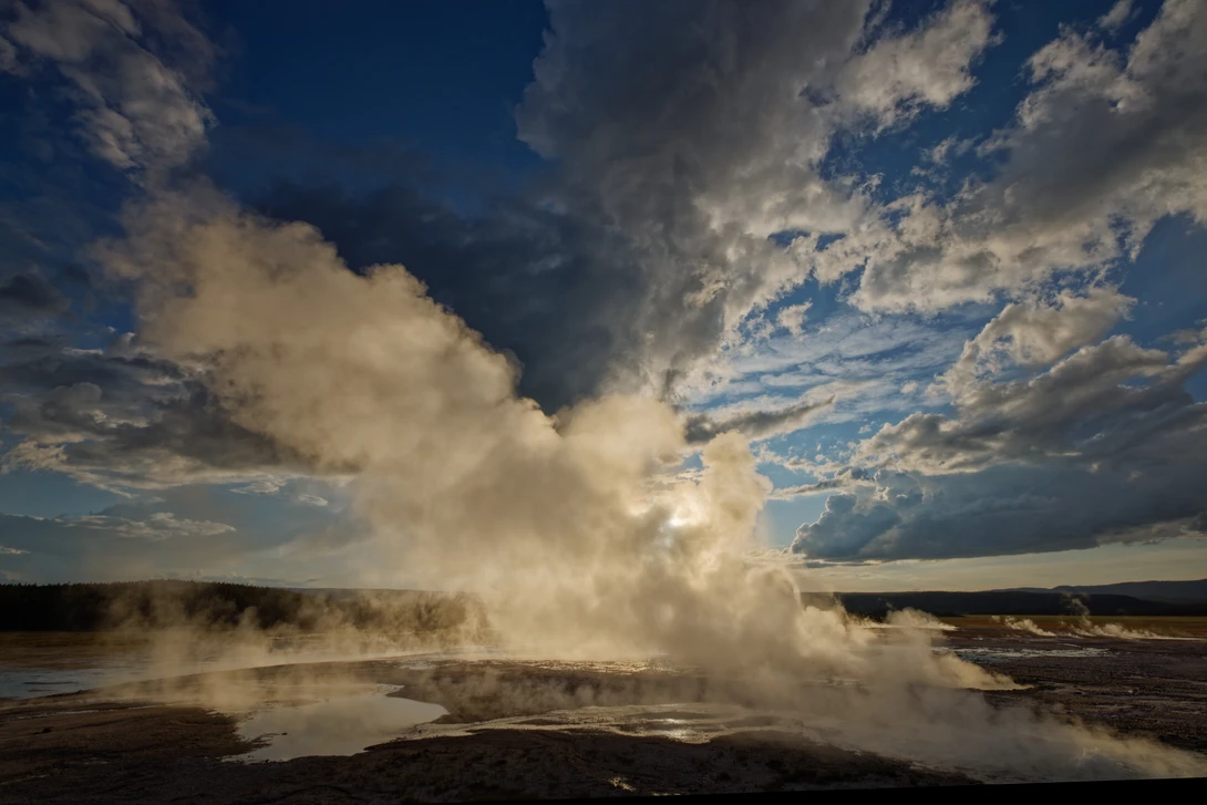 A geyser erupting a massive column of steam and hot water into a blue sky dotted with white clouds.