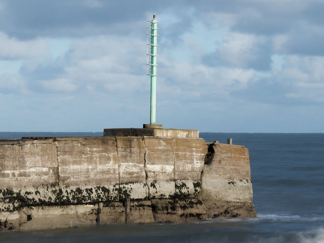 Weathered stone fort structure with tall green transmission tower rising from its center, surrounded by ocean and rocky shoreline.