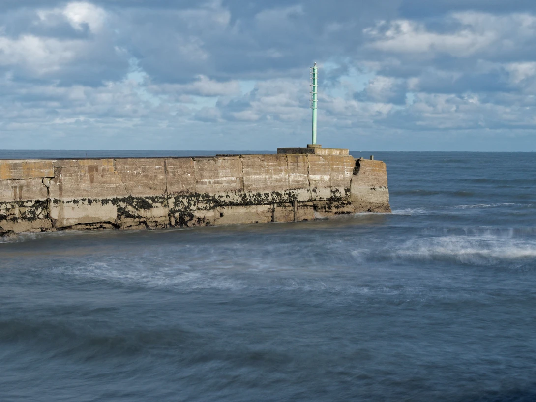 Stone pier extending into choppy ocean water under partly cloudy sky with lighthouse tower visible on the structure.