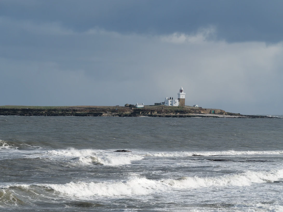 Coastal seascape with rolling waves and a distant lighthouse on a rocky headland under overcast sky.