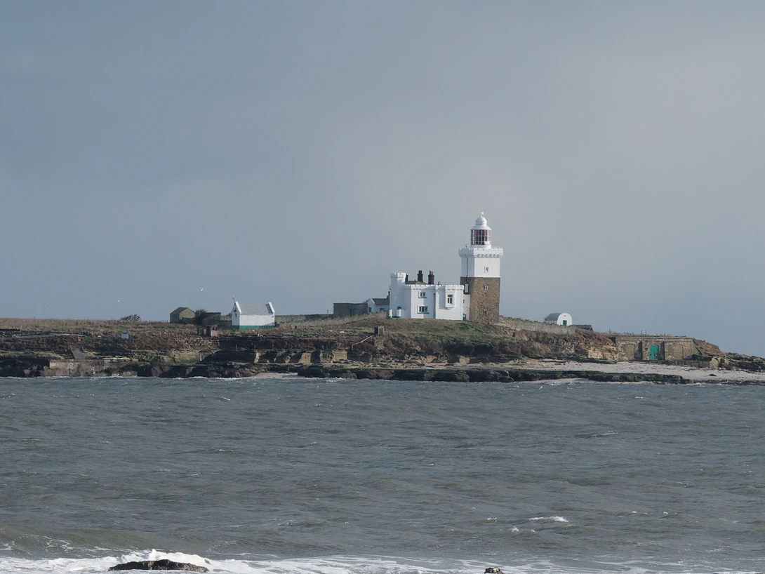 Coastal lighthouse and stone buildings on a rocky peninsula surrounded by choppy ocean water.