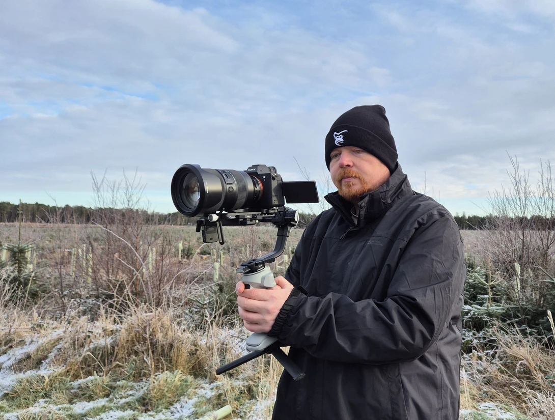 Filmmaker holding a cinema camera with telephoto lens in an open field during overcast daylight.