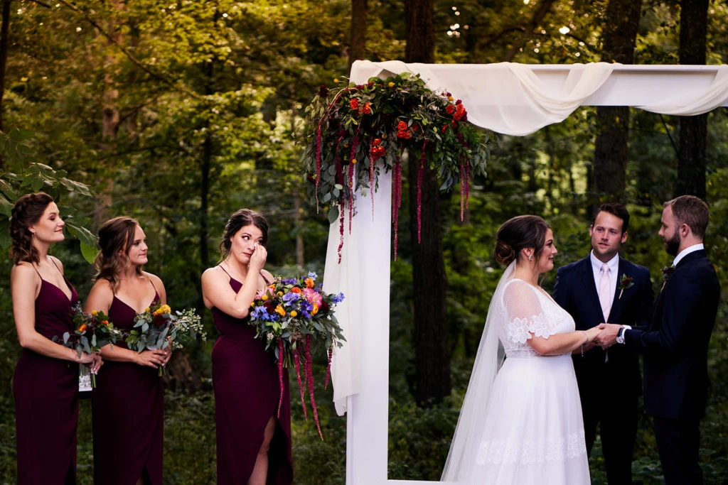 Bride and groom exchange vows at outdoor ceremony beneath white arbor decorated with cascading floral arrangements.