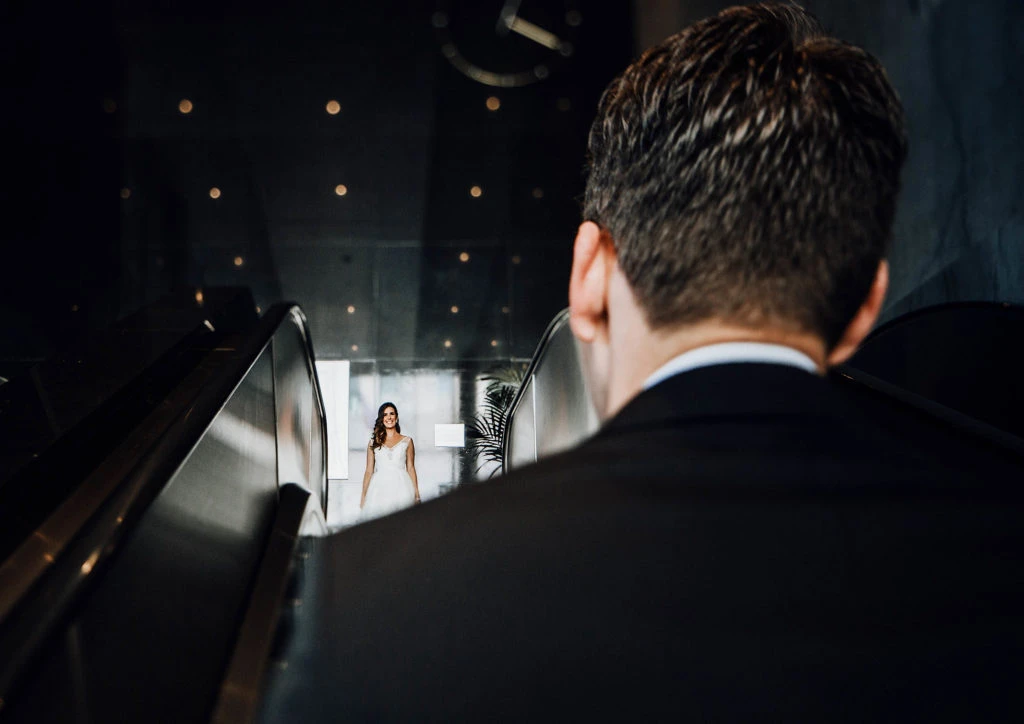 Man in dark suit watching a woman in white dress on large stage screen.