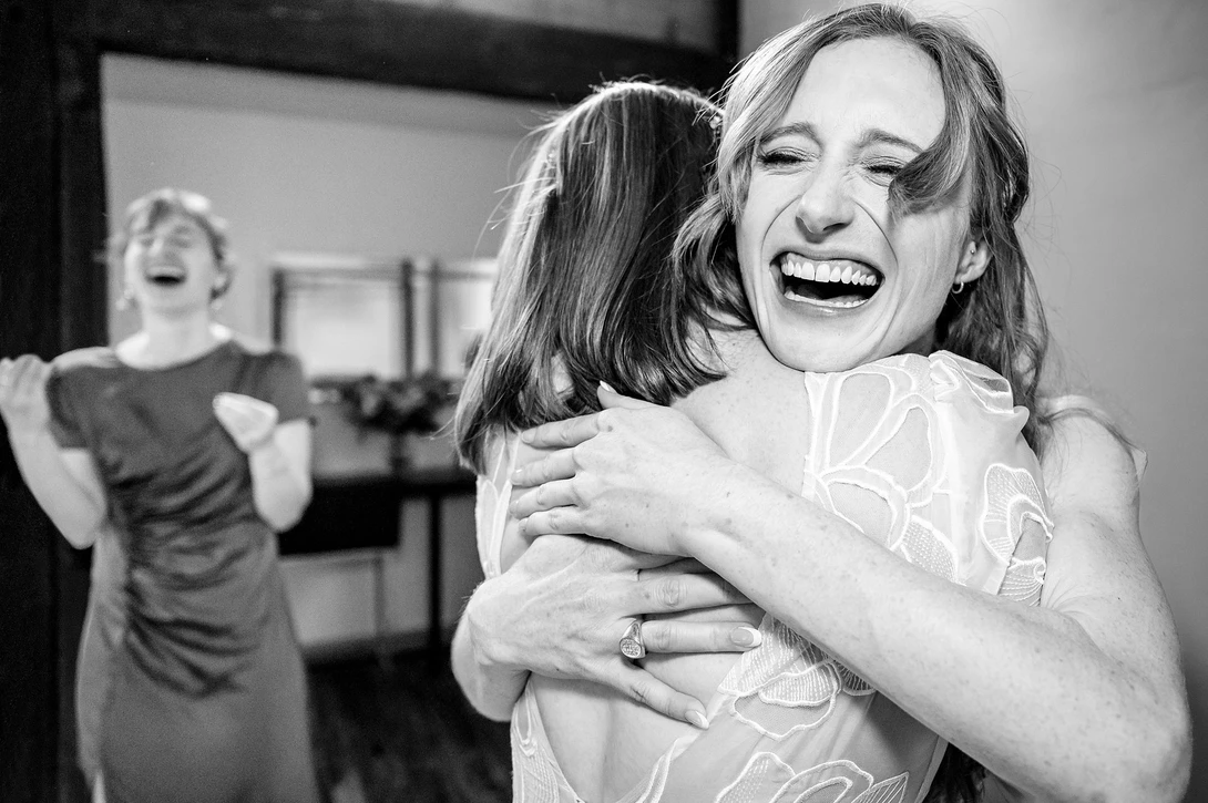 Woman laughing while hugging another person in an indoor setting with a third person visible in the background.