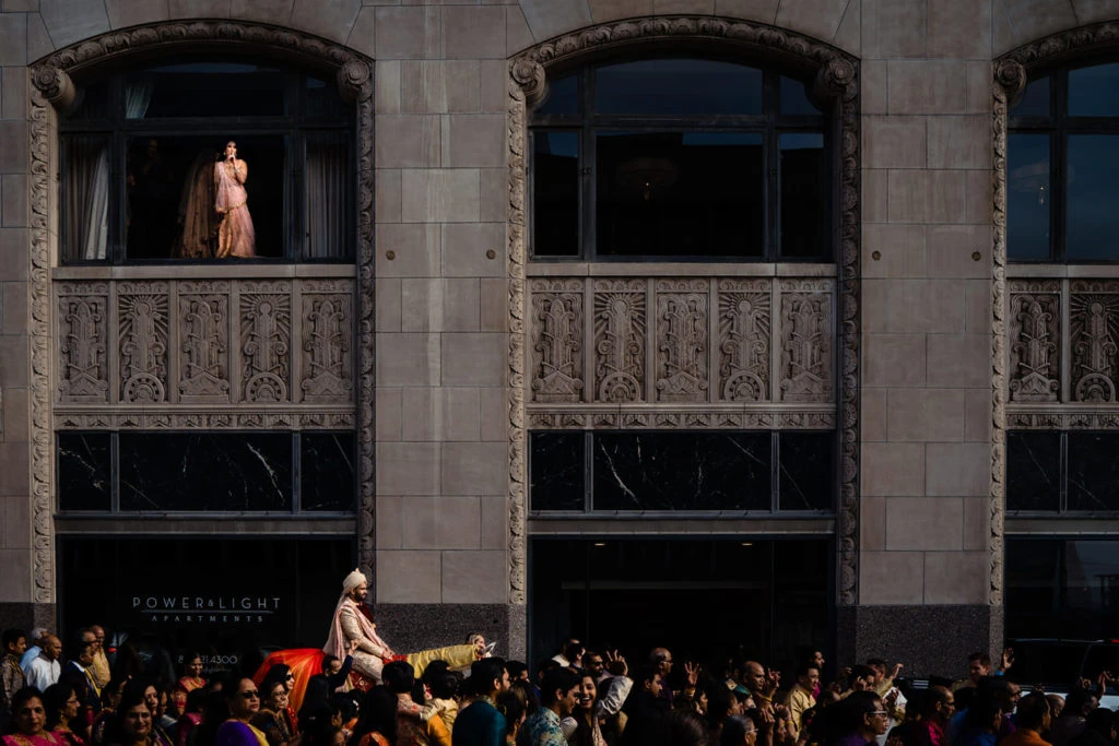 Fashion model on elevated runway in front of ornate arched windows with crowd gathered below.
