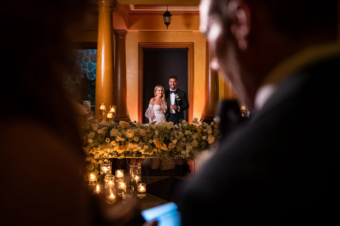 Bride and groom standing in a golden-lit doorway framed by flowering garland and hanging candles.