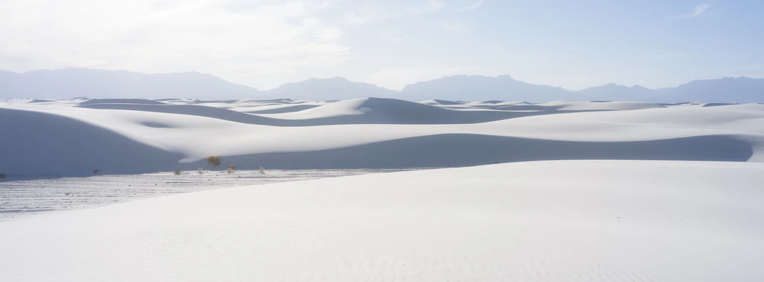 Undulating white gypsum dunes with mountains in the distance under hazy sky.