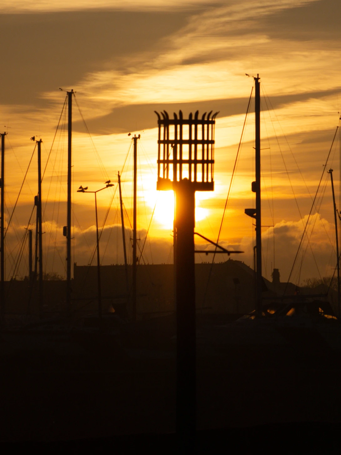 Silhouetted marina with sailboat masts and a tall wooden fire tower structure against a golden sunset sky.