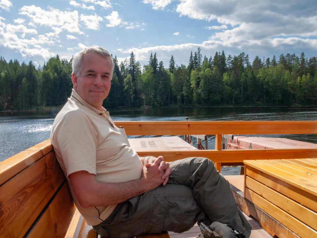 Man sitting on wooden boat deck with lake and coniferous forest in background.