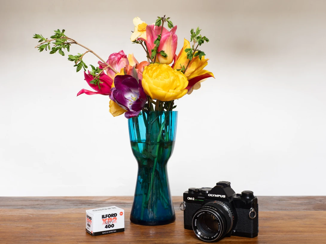 Colorful bouquet of ranunculus and roses in a teal glass vase, displayed with a vintage film camera and film roll.