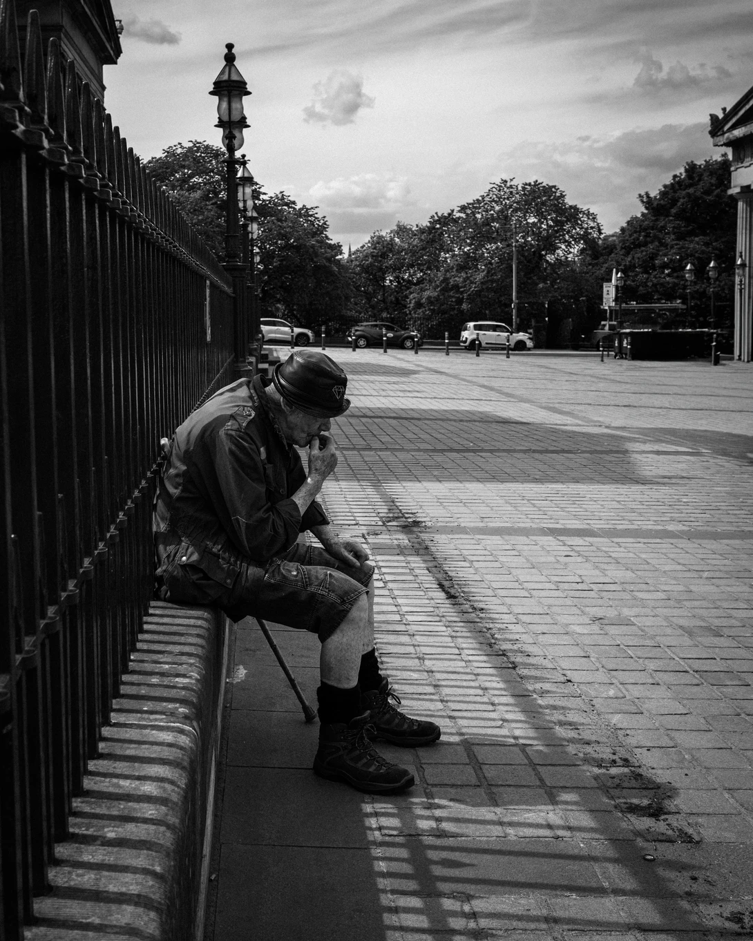 Person sitting on a bench against a metal fence in an urban plaza, rendered in black and white.
