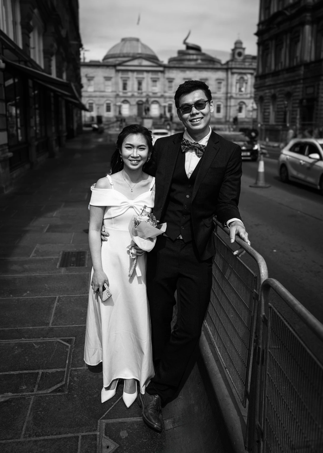 Couple in formal attire posing on a city street with classical architecture in the background.