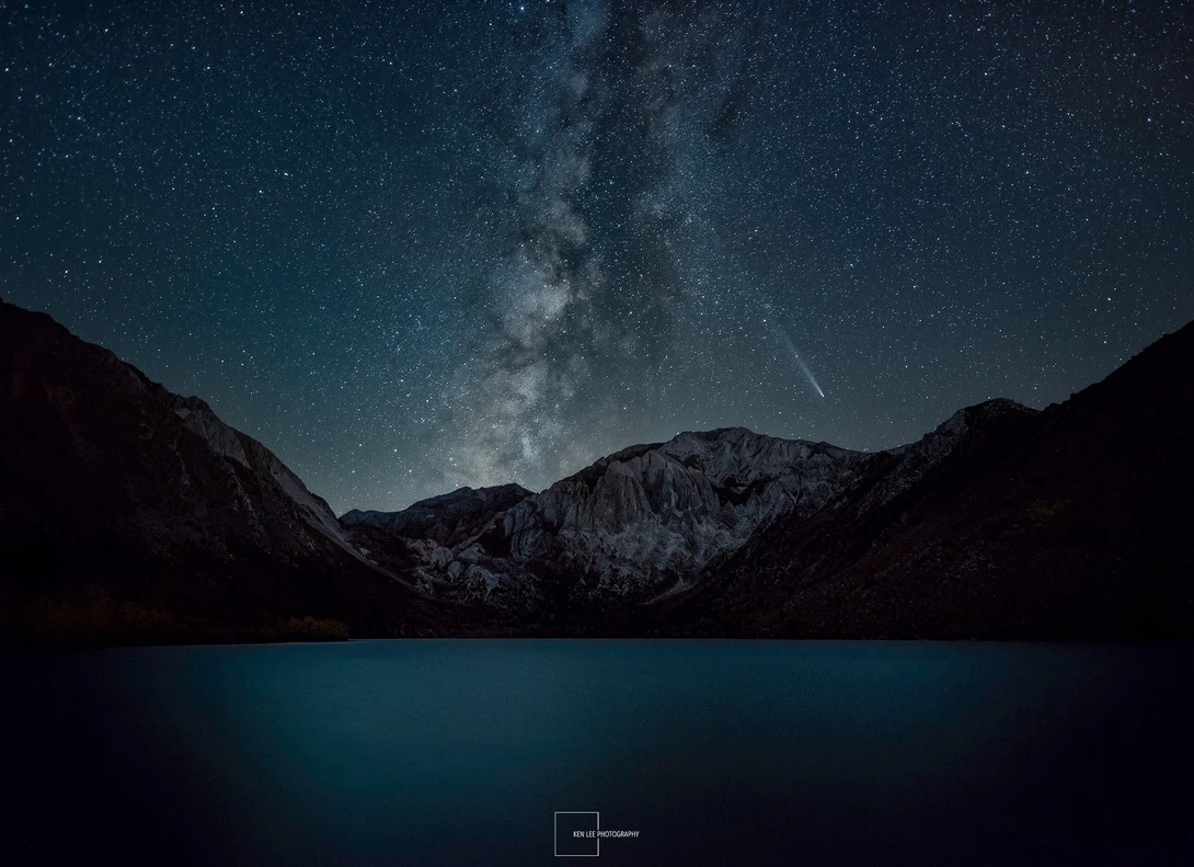 Milky Way galaxy arcing over a mountain lake at night with snow-capped peaks silhouetted against starry sky.
