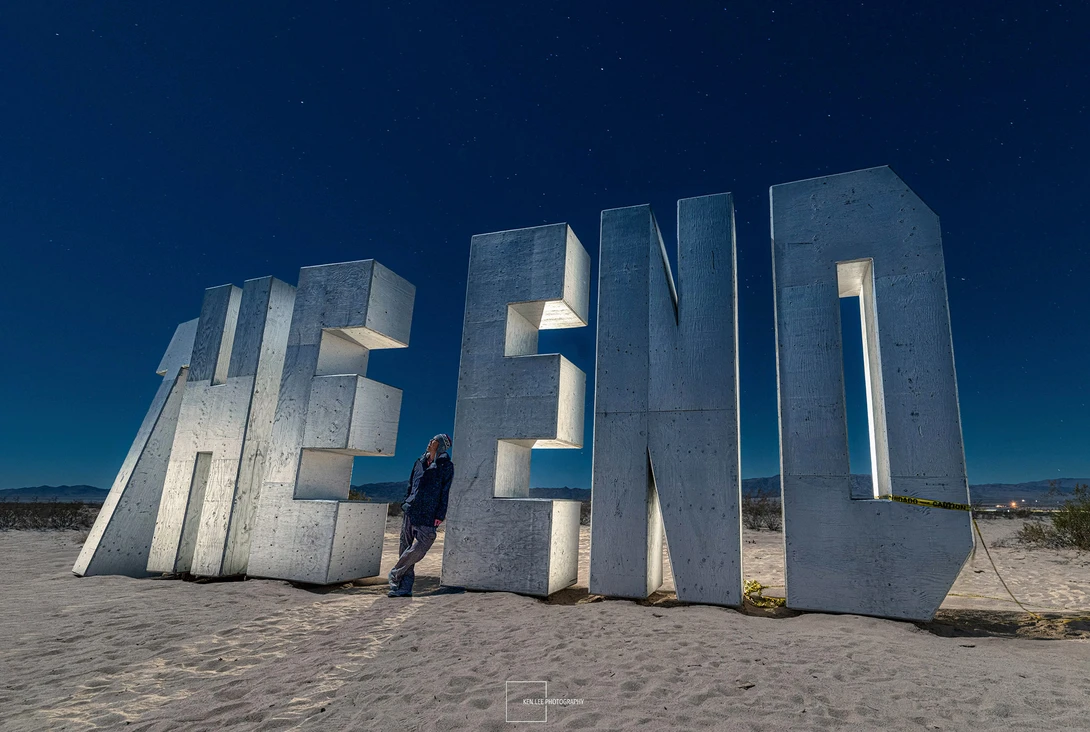 Person standing beside large concrete letter sculptures arranged on a sandy beach under clear blue sky.