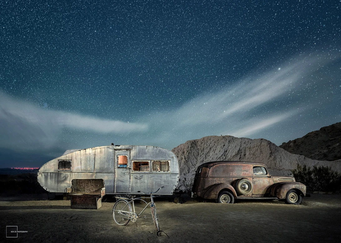 Vintage Airstream trailer and rusted car parked under a starry night sky with dramatic clouds.
