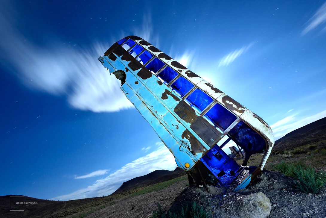 Blue and white bus tipped dramatically upward on a rocky outcrop against a bright blue sky.