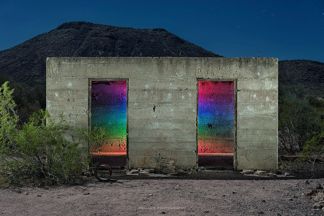 Concrete bunker with two doorways filled with vibrant rainbow-gradient light against desert mountain backdrop.