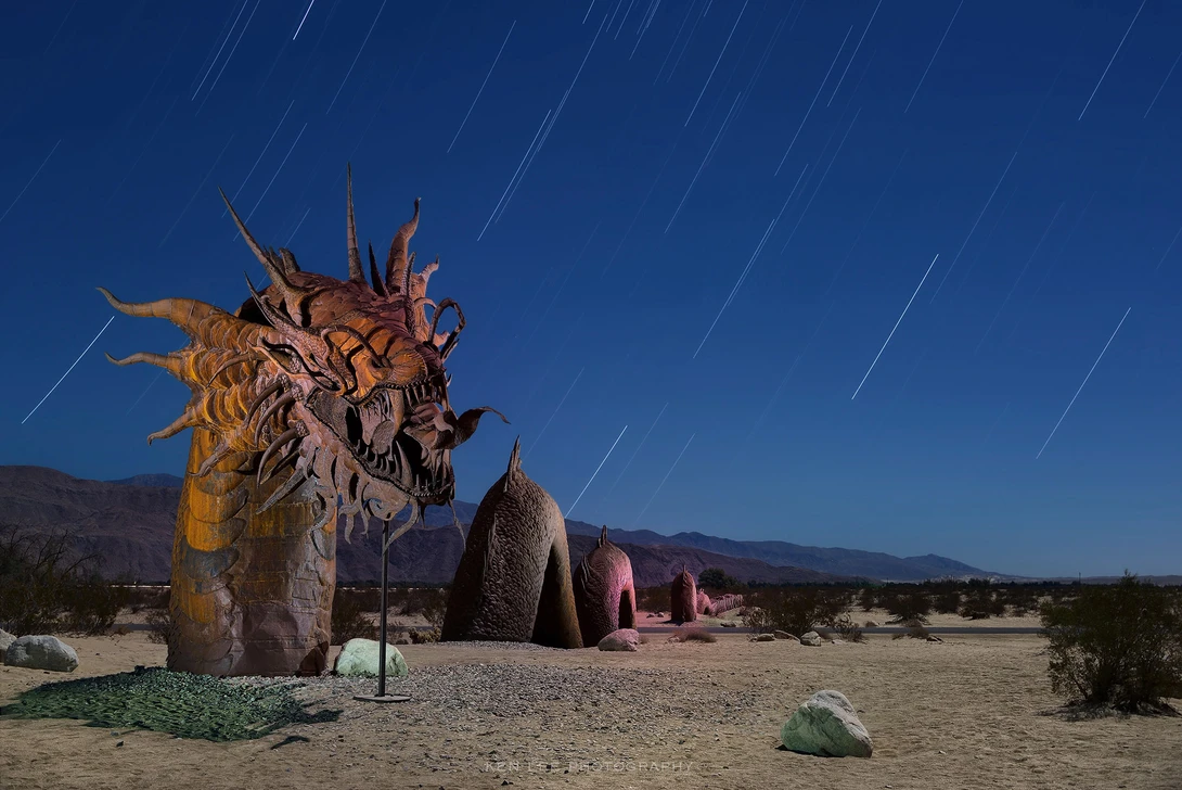 Desert landscape with sculptural rock formations and star trails streaking across deep blue night sky.