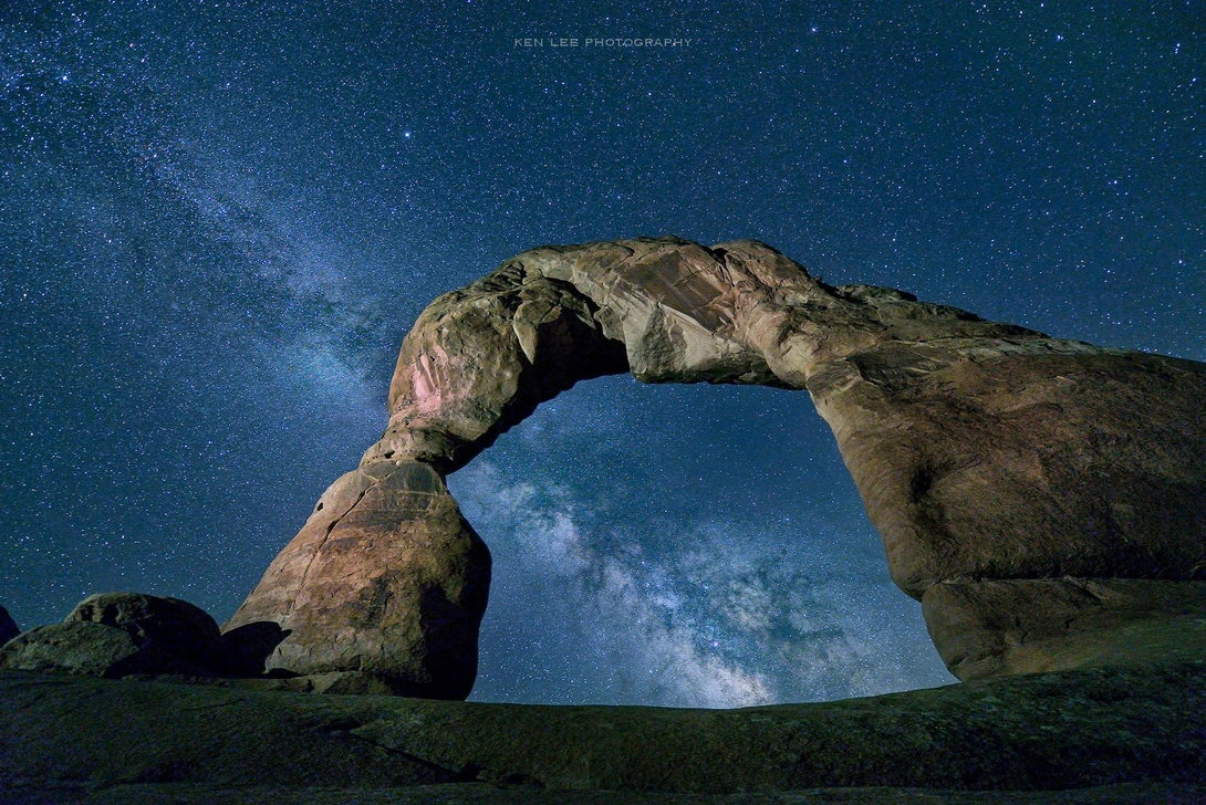 Natural stone arch framing the Milky Way galaxy against a dark blue night sky.
