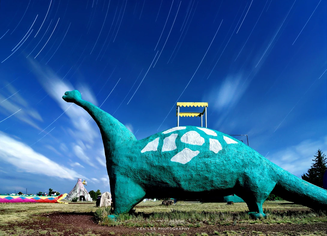 Oversized turquoise dinosaur sculpture with a small yellow structure on its back under a star-filled sky.