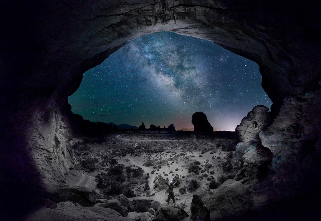 Silhouetted figure standing in a cave opening framing a starry night sky and Milky Way overhead.