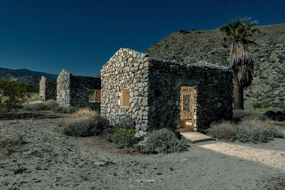 Stone structure with wooden door in desert landscape with palm tree and clear blue sky.