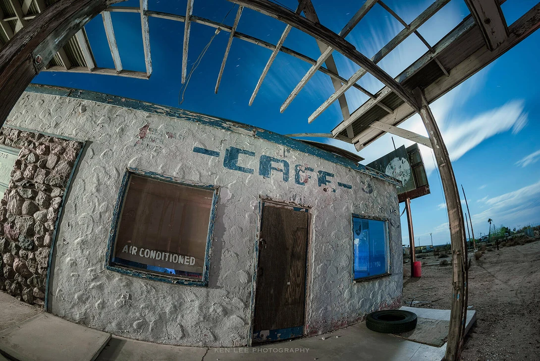 Abandoned concrete structure with metal frame roof photographed against clear blue sky using wide-angle lens.