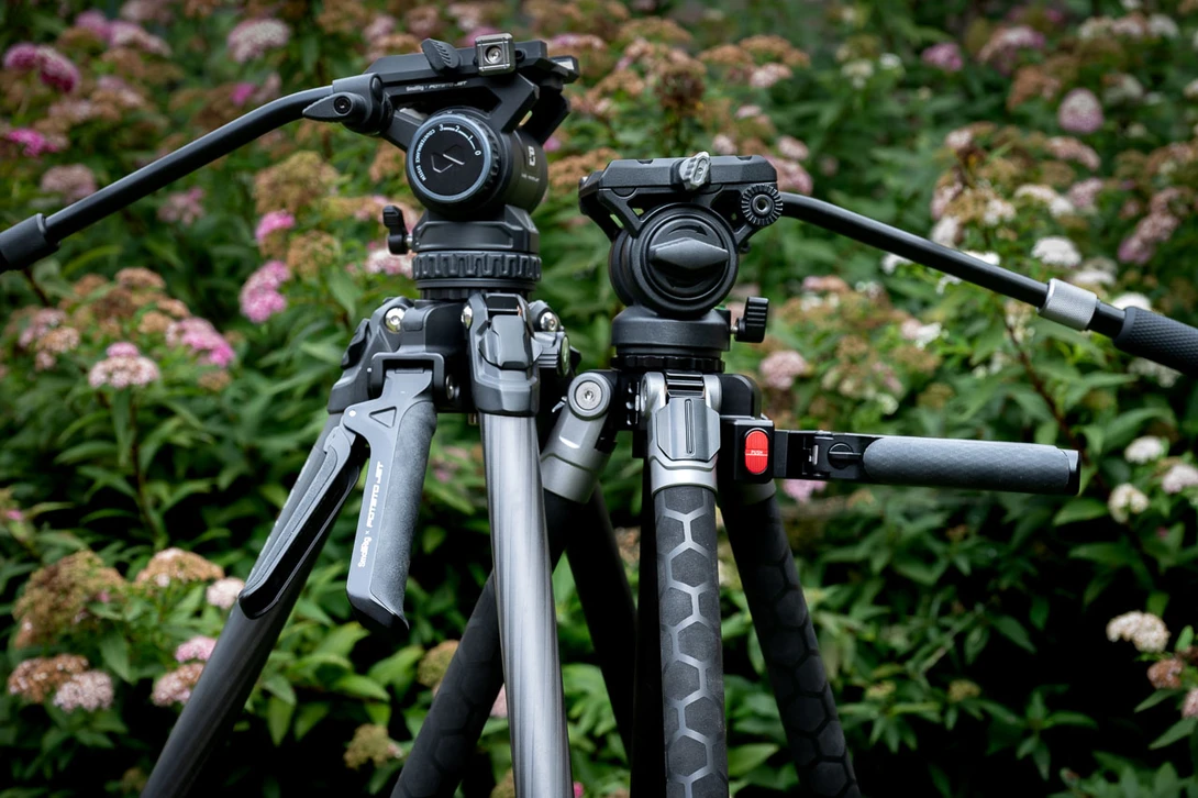 Two professional video tripods with fluid heads and pan handles positioned side by side against a blooming garden background.