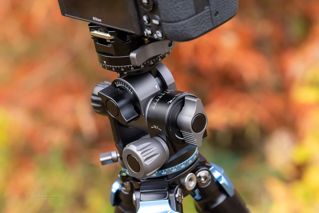 Professional camera tripod head with ball mount and geared pan-tilt mechanism against blurred autumn foliage background.
