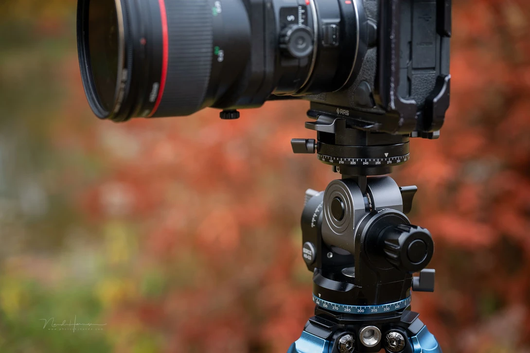 Camera with telephoto lens mounted on a geared tripod head with blurred autumn foliage background.