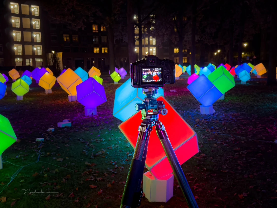 Nighttime urban plaza with glowing colorful cubic sculptures illuminated in vibrant neon colors, with camera on tripod in foreground.