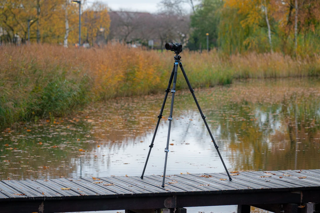 Camera on tripod positioned on wooden dock overlooking still water and autumn vegetation.