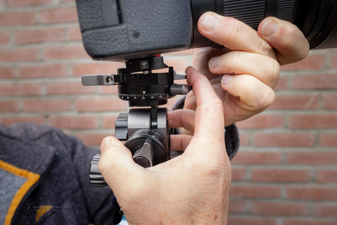 Hands holding a cinema camera with a mounted microphone and follow focus system against a brick wall backdrop.