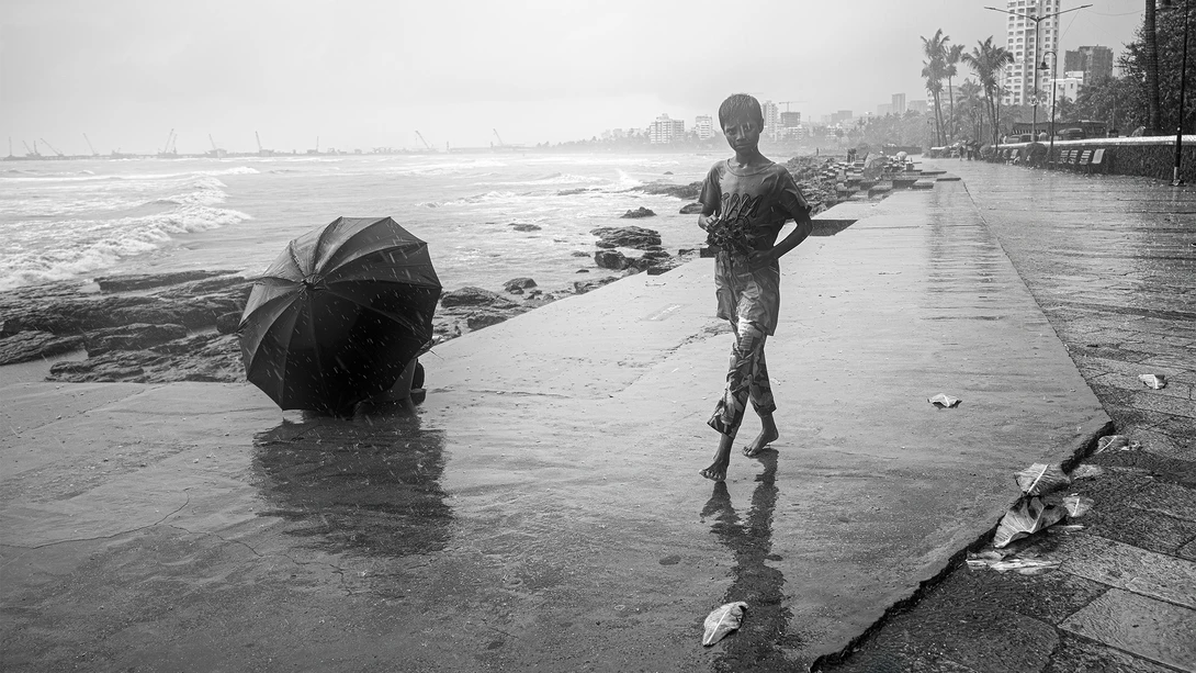 Person jogging on wet beach promenade during rain while another figure holds umbrella nearby.