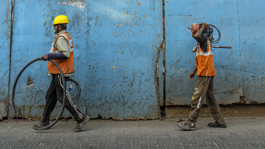 Two construction workers carrying bicycles and tools walk past a weathered blue metal wall.